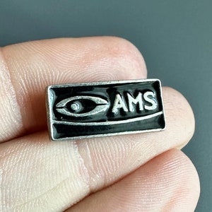 May include: A silver and black enamel pin with a stylized eye and the letters "AMS" in white.