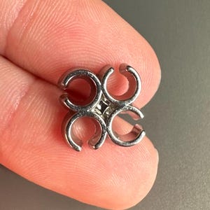 May include: A small, silver-coloured metal charm with a four-leaf clover design. The charm is composed of four curved, open shapes joined at the centre. The charm is held between two fingers against a grey background.