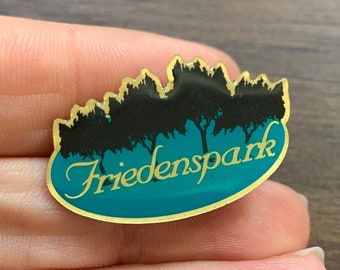 May include: A gold-colored pin with a teal blue background and a black silhouette of trees. The text "Friedenspark" is written in gold lettering on the pin.
