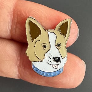 May include: A corgi enamel pin featuring a detailed illustration of a corgi's head. The pin has a tan and white face with a blue collar. The corgi's ears are tan, and its tongue is sticking out.