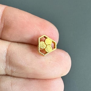 Puede incluir: Un colgante hexagonal de color dorado con un diseño de panal de abeja. El colgante presenta un patrón en relieve de cuatro círculos dentro del hexágono.