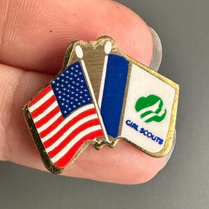 May include: A gold-coloured pin depicting two flags. One is the American flag, and the other has a blue background with a white emblem and the words "GIRL SCOUTS". The pin is small and likely made of metal.