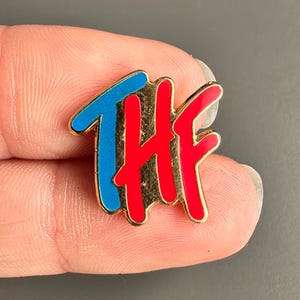 May include: A gold-toned pin with the letters "THF" in a stylized script. The "T" is blue, and the "HF" is red, with a gold outline. The pin is small and appears to be enamel-filled.