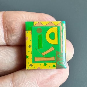May include: A green and yellow enamel pin with the word "id" in a stylized font. The pin has a colorful geometric design with stars and wavy lines.