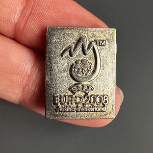 Puede incluir: Un pin rectangular plateado con el logotipo EURO 2008. El pin tiene el texto "EURO 2008" y "Austria-Suiza" en una fuente estilizada. El pin también tiene un logotipo con las letras "my" y un diseño estilizado.