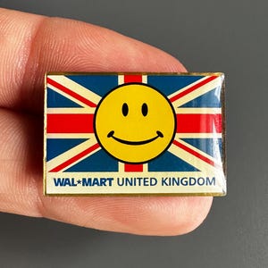 May include: A rectangular pin featuring the Union Jack flag design with a yellow smiley face in the center. The pin has the text "WAL*MART UNITED KINGDOM" at the bottom. The pin is held against a gray background.
