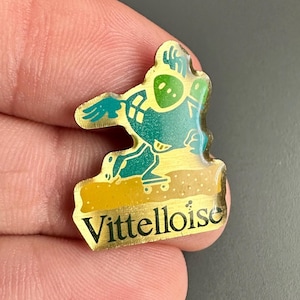 May include: A gold-colored pin with a cartoon character riding a skateboard. The character is green and yellow and has a large head. The pin says "Vitteltoise" in gold letters.