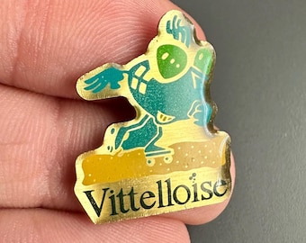 Könnte beinhalten: Eine goldfarbene Anstecknadel mit einer Comicfigur auf einem Skateboard. Die Figur ist grün und gelb und hat einen großen Kopf. Die Anstecknadel trägt die Aufschrift "Vitteltoise" in goldenen Buchstaben.