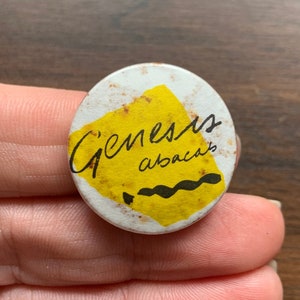 May include: A white button with a yellow and black design. The button says "Genesis abacus" in black lettering.