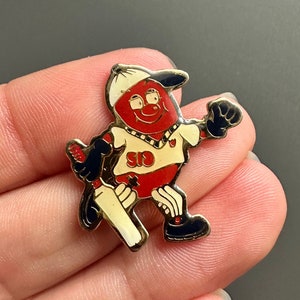 Könnte beinhalten: Eine Emaillestifte mit einem Cartoon-Charakter, der einen Cricket-Schläger hält. Der Charakter trägt ein weißes Hemd mit dem Wort "SID" darauf, eine rote Mütze und schwarze Schuhe.