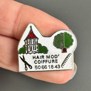 May include: A small, enamel pin with a white and red building, a green tree, and the text "HAIR MOD' COIFFURE 50 66 18 43". The pin also features a pair of scissors and a comb.
