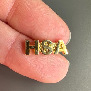 May include: A small, gold-colored pin with the letters "HSA" in capital letters. The pin is held between two fingers against a gray background. The pin appears to be made of metal and has a shiny finish.