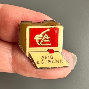Könnte beinhalten: Eine goldfarbene Anstecknadel in Form eines Computerbildschirms mit einem roten Quadrat, das ein stilisiertes Euro-Symbol zeigt. Die Nadel hat den Text "3616 ECUBANK" unten. Die Nadel wird in der Hand einer Person gehalten.
