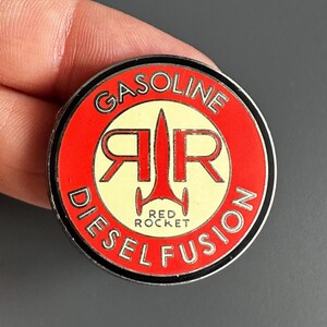 Peut inclure: Un pin's circulaire en émail avec une bordure rouge portant le texte blanc GASOLINE DIESEL FUSION. Le centre est crème avec un motif de fusée rouge entouré des lettres R et R, et le texte RED ROCKET imprimé sous la fusée.