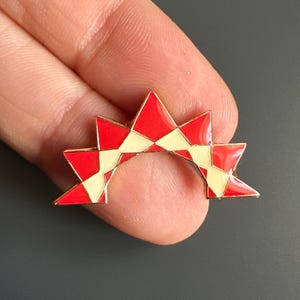 May include: A red and cream-colored enamel pin in a starburst design. The pin features a series of red and cream-colored triangles arranged in a star-like pattern. The pin is held in a person's hand against a dark gray background.