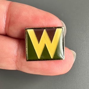 Puede incluir: Un pin cuadrado con borde dorado. El pin presenta una parte superior e inferior marrón oscuro, con una sección media verde. Una "W" amarilla está centrada en el pin. El pin está sostenido por los dedos de una persona.