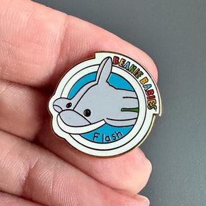 May include: A gold-coloured pin featuring a white dolphin design and the text "Beanie Babies" in a rainbow colour scheme. The dolphin is surrounded by a blue circle with the word "Flash" in white.