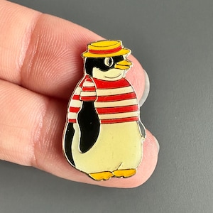 May include: A colorful penguin pin with a yellow belly, black wings, and a red and white striped shirt. The penguin wears a yellow hat with a black band. The pin is held in a hand against a gray background.