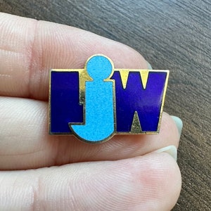 May include: A gold-colored pin with the letters "IJW" in blue and purple enamel. The "J" is a stylized figure of a person.