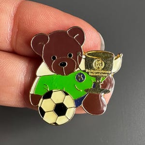 May include: A brown teddy bear pin wearing a green shirt and blue pants, holding a soccer ball and a gold trophy. The soccer ball has black and white pentagons. The trophy has a gold finish and the letter "K".