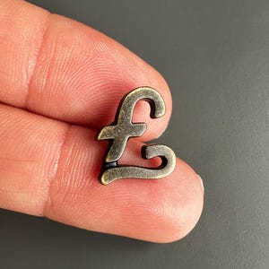 May include: A small, metallic, antique-bronze colored charm shaped like the British pound sterling currency symbol. The charm has a smooth, curved design with a dark, weathered finish and is held between two fingers for scale.