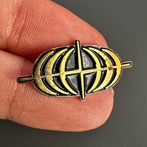 May include: A small, oval-shaped pin with a gold-colored globe design, featuring black accents and a cross-shaped element. The pin has a dark border and is held between fingers against a dark background. The pin is approximately 1 inch in length.