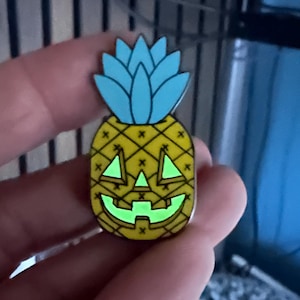 May include: A metal enamel pin shaped like a pineapple with a blue top and a yellow body. The pineapple features a carved jack-o'-lantern face with glowing green eyes and a mouth, accented by small black cross patterns across the yellow surface.