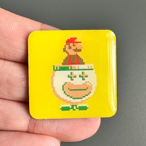 May include: A yellow square pin featuring a pixelated image of a video game character. The character is wearing a red hat and is inside a white cup with a green bow tie and a smiling face.