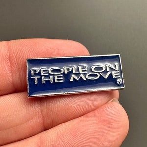 May include: A blue enamel pin with silver trim that reads "PEOPLE ON THE MOVE" with a registered trademark symbol.