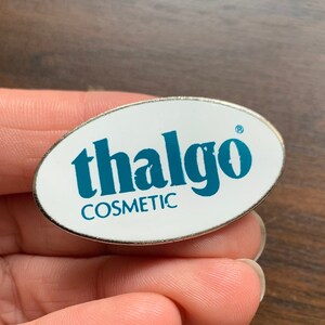 Puede incluir: Un pin ovalado blanco con un logotipo azul que dice "thalgo COSMETIC" y un símbolo de marca registrada.