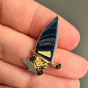 Puede incluir: Un pin de color dorado con un diseño de velero azul. El velero tiene el texto "GAULOISES BLONDES".