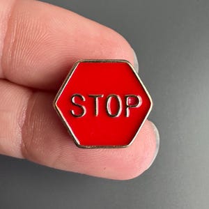 May include: A hexagonal enamel pin featuring a bright red background with the word STOP embossed in gold-toned metal lettering. The pin has a thin gold-toned metal border outlining the hexagonal shape, creating a clear and high-contrast design.