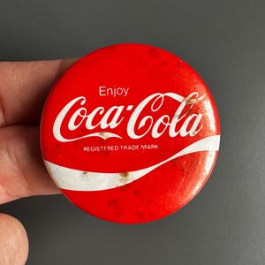 May include: A round, red Coca-Cola button with the Coca-Cola logo in white script. The button also includes the words "Enjoy" and "REGISTERED TRADE MARK".