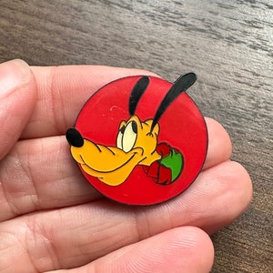 May include: A round pin featuring Pluto, a yellow dog with black ears and a black nose, set against a red background. Pluto is holding a green and red object. The pin is held in a hand.