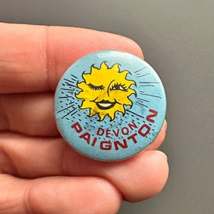 Könnte beinhalten: Ein blauer und gelber Knopf mit einem lächelnden Sonnen-Gesicht und dem Text "Devon Paignton" in Rot.
