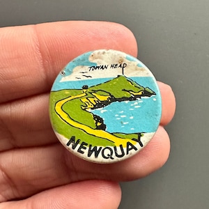 Puede incluir: Un botón redondo blanco con una ilustración colorida de una colina verde con un faro en la cima. El texto "Towan Head" y "Newquay" está impreso en el botón.