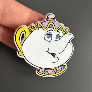 Op de afbeelding: Een witte theepot in cartoonstijl met een lachend gezicht, blauwe ogen en een paarse en gele deksel. De theepot heeft een gele handgreep en tuit, en een decoratieve kraag met roze, gele en blauwe accenten. Het ontwerp is zwart omlijnd.