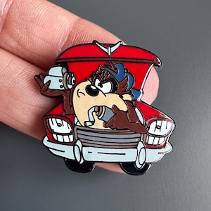 Op de afbeelding: Een kleurrijke emaille pin met de Tasmanian Devil uit Looney Tunes in een rode auto. De pin heeft een gedetailleerd ontwerp met zwarte, bruine, witte en blauwe accenten. De motorkap van de auto staat open en het personage draagt een blauwe pet.