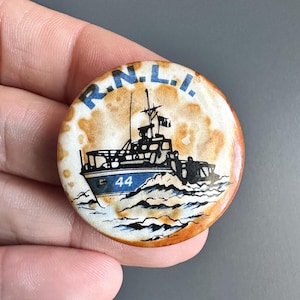 Puede incluir: Un botón redondo blanco con una pátina marrón oxidada. El botón presenta una ilustración en blanco y negro de un bote salvavidas con el texto "R.N.L.I." y el número "44" en el lateral.