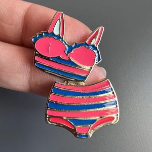 May include: A metal enamel pin shaped like a two-piece swimsuit. The bikini top and high-waisted bottoms feature horizontal stripes in vibrant pink and royal blue, outlined with gold-toned metal edges. The design includes small white accents.