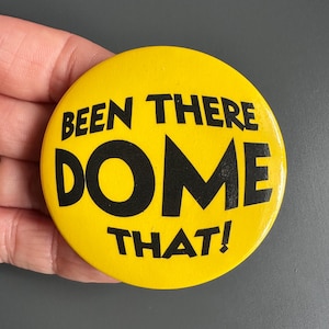 以下が含まれることがあります： 「BEEN THERE DOME THAT!」という黒い太字のテキストが書かれた、丸い黄色のボタン。ボタンは灰色の背景に対して手で持たれています。