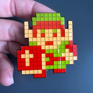 May include: A pixel art enamel pin featuring a character with a green hat, tan face, and red tunic, holding a large red shield with a yellow cross design. The figure is constructed from a grid of small squares in shades of green, red, and tan.
