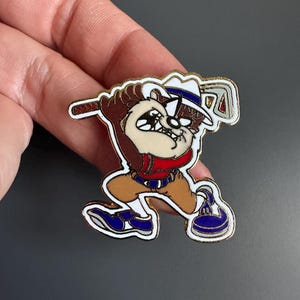 May include: A colourful enamel pin featuring the Tasmanian Devil cartoon character playing golf. The pin has a gold border and depicts Taz in a golf outfit, swinging a golf club. The design includes shades of brown, red, blue, and white.