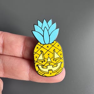 Puede incluir: Un pin de esmalte metálico con forma de piña, cuerpo amarillo brillante y hojas azul claro. La piña presenta una cara de calabaza tallada con ojos triangulares y una amplia sonrisa dentada, acentuada por pequeños detalles de cruces negras.