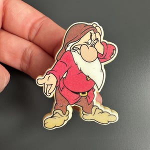 Può includere: Immagine di un cartone animato di Brontolo di Biancaneve. Il personaggio indossa una camicia rossa, pantaloni marroni e un cappello marrone. Ha una barba bianca e si tocca la fronte con la mano.