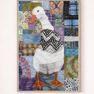 Könnte beinhalten: Gerahmtes Kunstwerk mit einer weißen Gans mit orangefarbenem Schnabel und Füßen, die einen schwarz-weiß gemusterten Pullover trägt. Der Hintergrund ist ein Patchwork aus verschiedenen Stoffmustern in Blau-, Grün-, Lila- und Brauntönen.