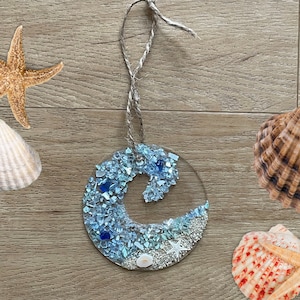 May include: A blue and white wave-shaped ornament made of resin with embedded seashells and glass. The ornament is hanging from a twine string.