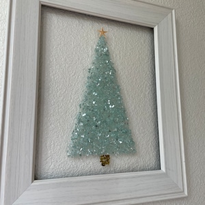 May include: A white framed picture with a light blue glass Christmas tree inside. The tree is made of small pieces of glass and has a gold star on top.