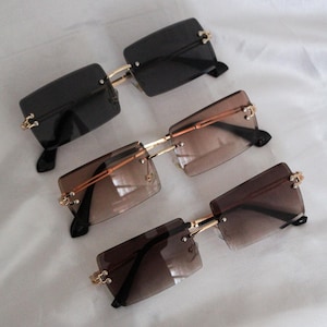 May include: Three pairs of square-shaped sunglasses with gold frames. The sunglasses have different coloured lenses: black, brown, and a gradient of brown.