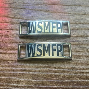 Puede incluir: Dos abalorios de zapatos plateados con el texto "WSMFP" en letras negras.